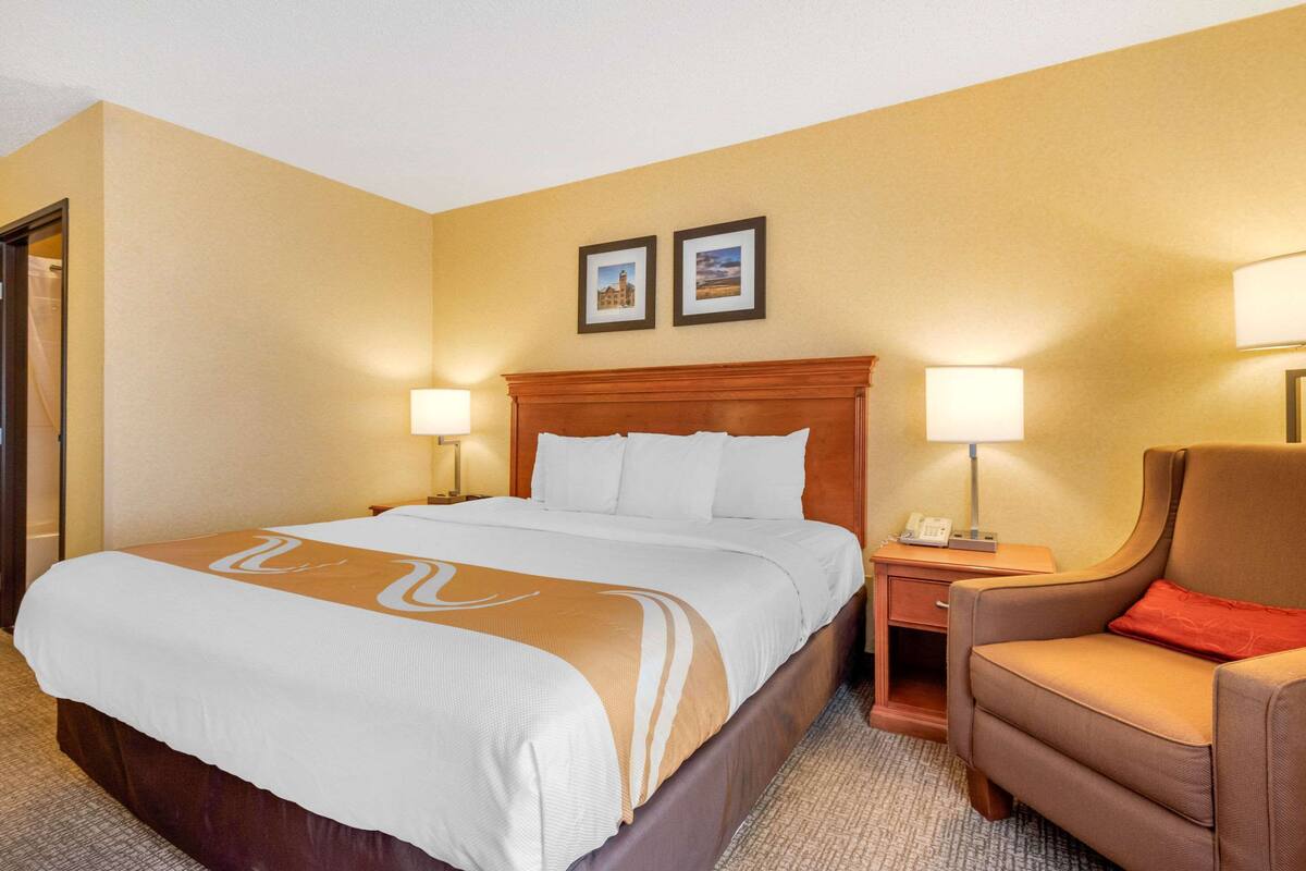 A hotel room with a large bed, two bedside lamps, a wooden headboard, a chair, and two framed pictures on the wall.
