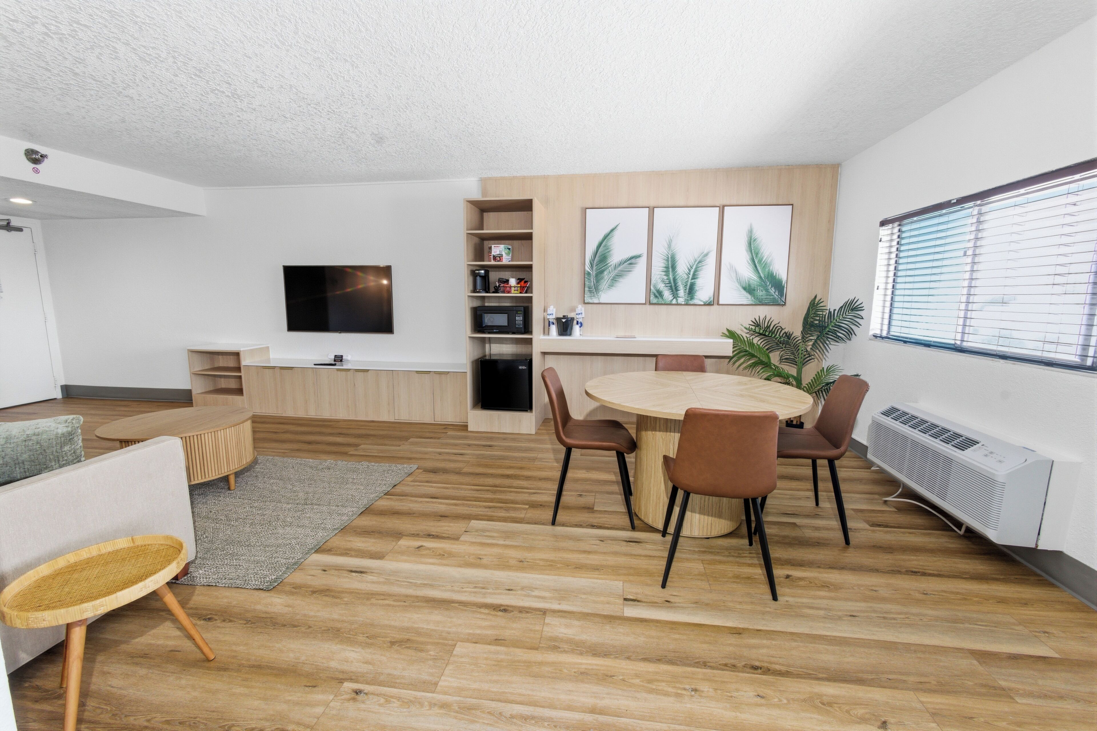 A modern living room with a round dining table, chairs, a flat-screen TV, a built-in kitchen area, and a large window with blinds.