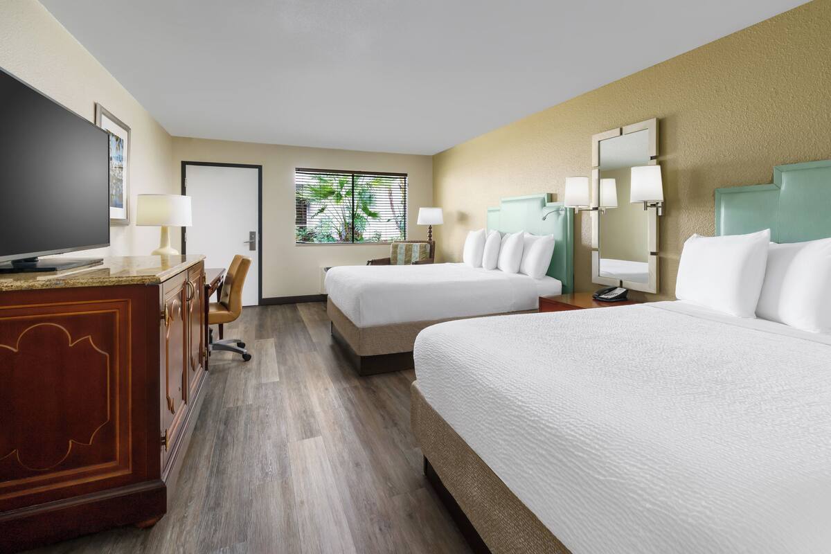 Quarto de hotel com duas camas, televisão, escrivaninha com luminária e janela com vista para o verde.