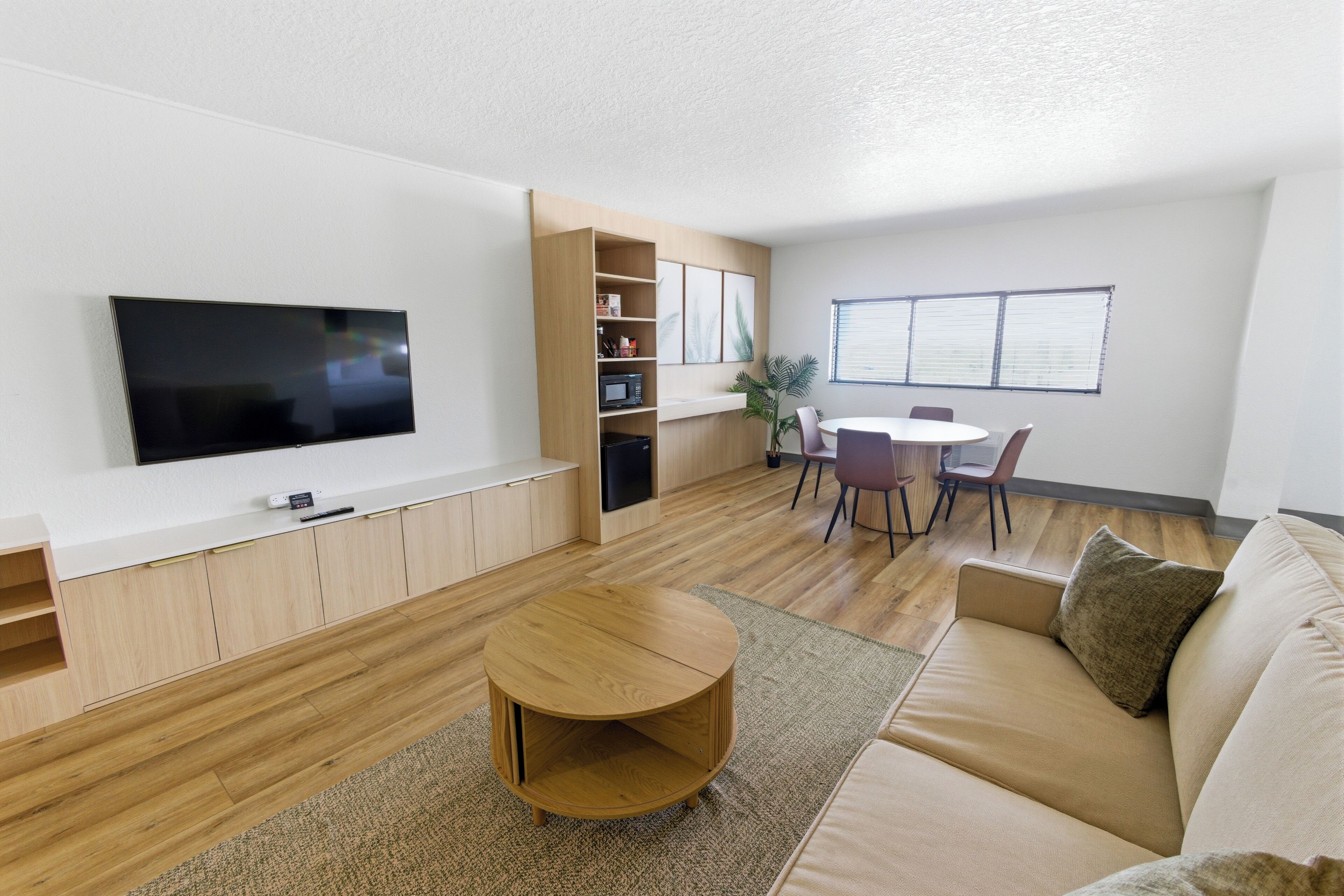 A modern living room with a flat-screen TV, wooden shelves, a round coffee table, a sofa, and a dining area with chairs and a table.