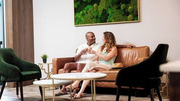 A couple sitting on a leather couch in a well-lit room with a green chair, a small round table, and a wall-mounted plant.