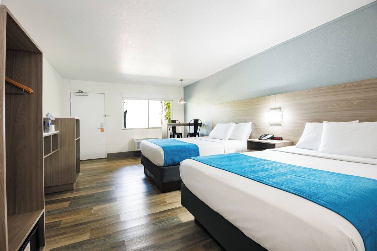 Quarto de hotel com duas camas, piso de madeira, guarda-roupa e uma escrivaninha com cadeira.