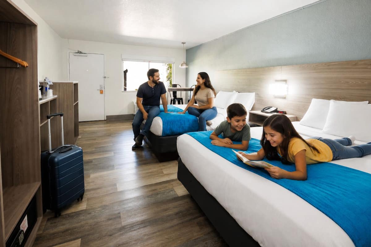 Quarto de hotel com duas camas, um guarda-roupa de madeira, uma mala e uma escrivaninha com uma cadeira.