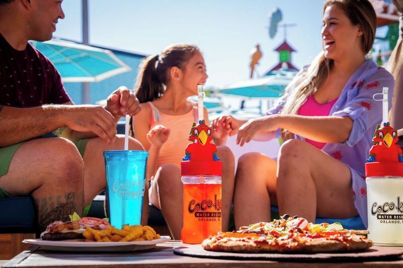 Un groupe de personnes est assis autour d’une table, devant des plats et des boissons, notamment un hamburger, des frites et une pizza, ainsi que des bouteilles d’eau de la marque Coca-Cola.