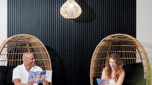 Two people seated in wicker chairs, reading magazines, with a black wall and a hanging light fixture in the background. - Coco Key Hotel & Water Park Resort (Orlando)