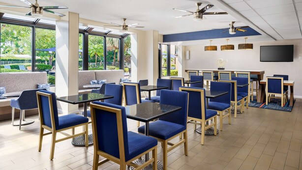 Un comedor moderno con sillas tapizadas en azul y mesas, ventanas amplias, ventiladores de techo y un televisor de pantalla plana en la pared.