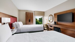 Habitación de hotel con dos camas, un escritorio, un televisor y una ventana con vistas a árboles.