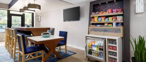 Un comedor moderno con una mesa de madera y sillas azules, un mostrador de aperitivos con diferentes opciones, un refrigerador para bebidas y un televisor montado en la pared.