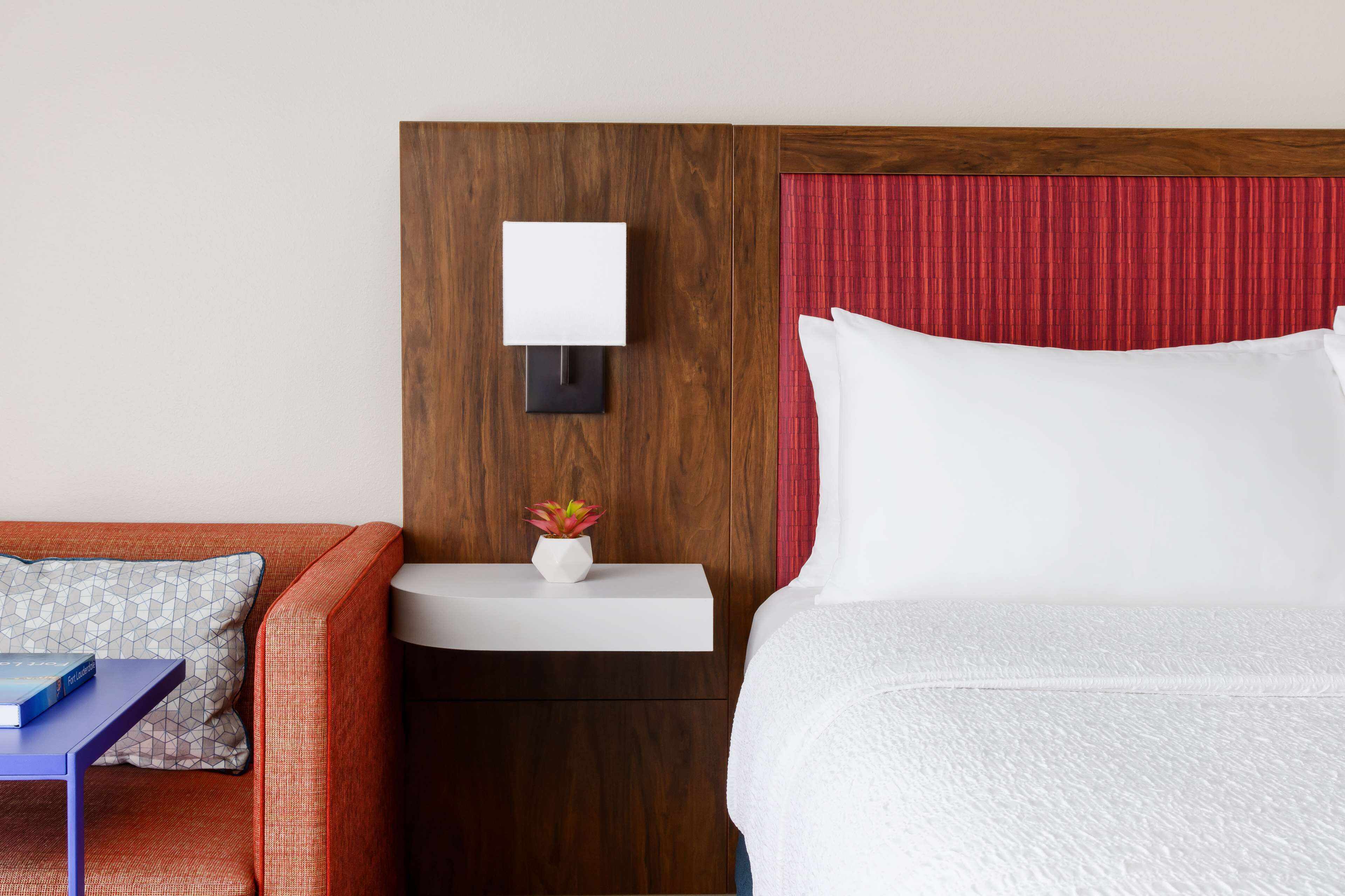 Habitación de hotel con cabecera de madera, mesita de noche blanca con lámpara, cortina roja, cama blanca, sofá rojo con cojines estampados y una pequeña planta en maceta.