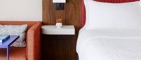 Habitación de hotel con cabecera de madera, mesita de noche blanca con lámpara, cortina roja, cama blanca, sofá rojo con cojines estampados y una pequeña planta en maceta.