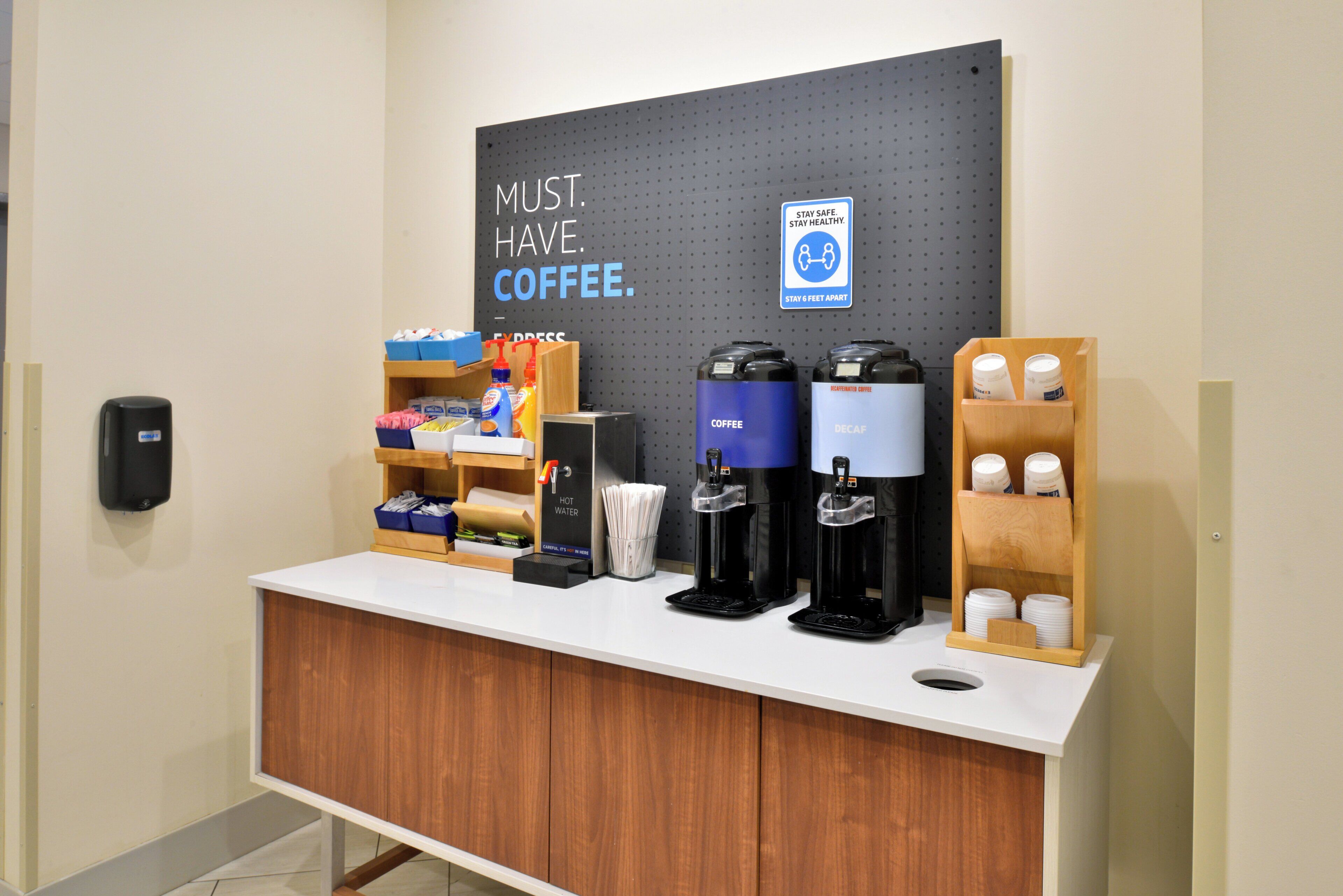 A coffee station with dispensers, a sign that says 'MUST HAVE COFFEE,' and a 'STAY SAFE. STAY HEALTHY. STAY 6 FEET APART' sign.