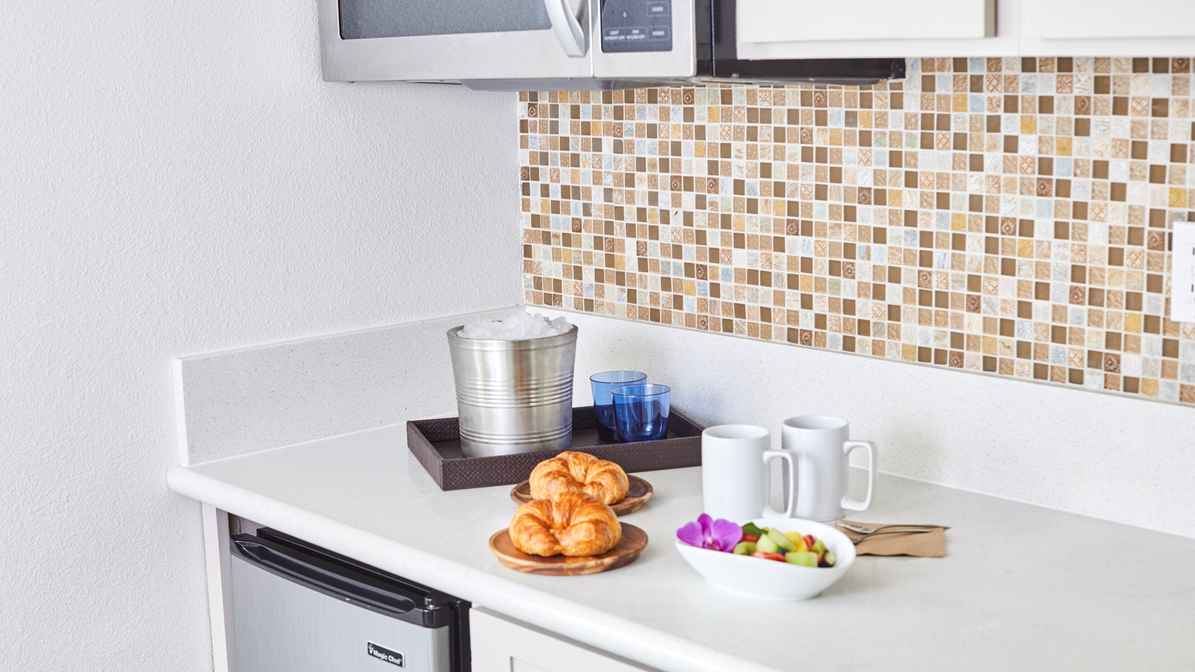Una kitchenette con microondas, cafetera, croissants y un bol de fruta.