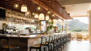 A bar with a long counter, multiple hanging pendant lights, and a view of mountains. - Queen Kapiolani Hotel (Honolulu)