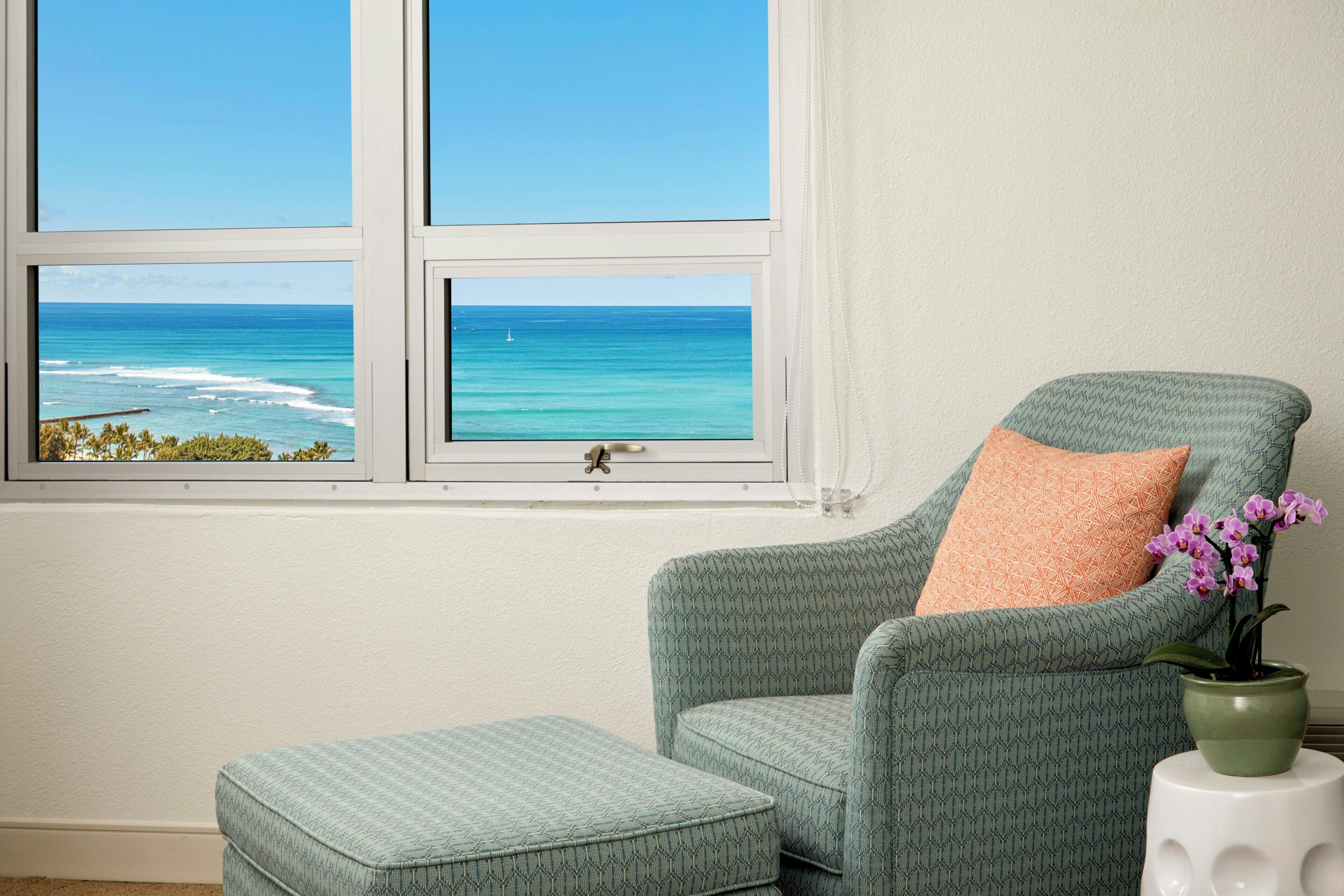 Une pièce avec vue sur l’océan, visible par une fenêtre, comprenant un fauteuil à motifs turquoise, un pouf assorti et une orchidée en pot posée sur un tabouret blanc.