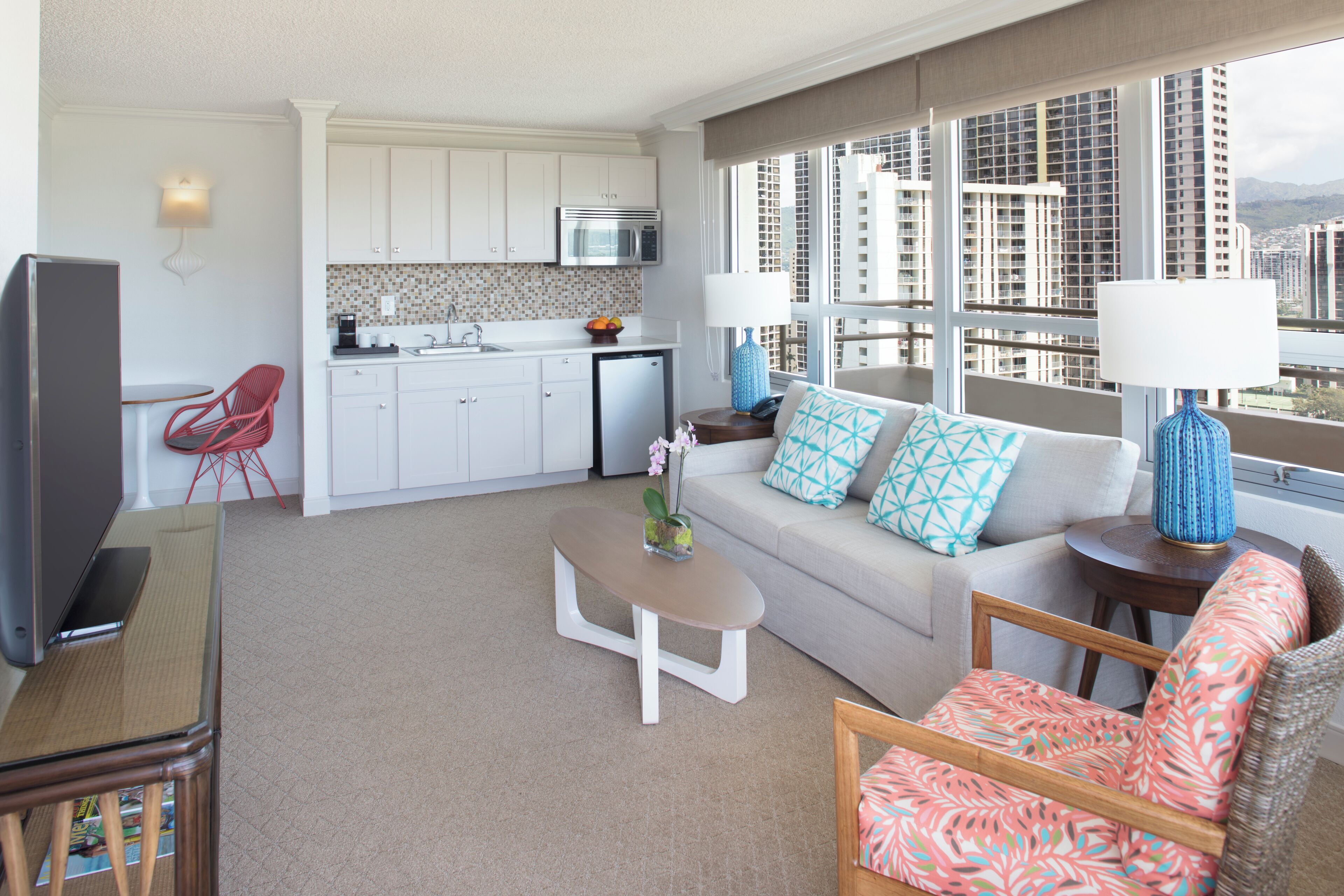 A modern living room with a sofa, coffee table, and red chair. A kitchen with white cabinets and a microwave is visible in the background.