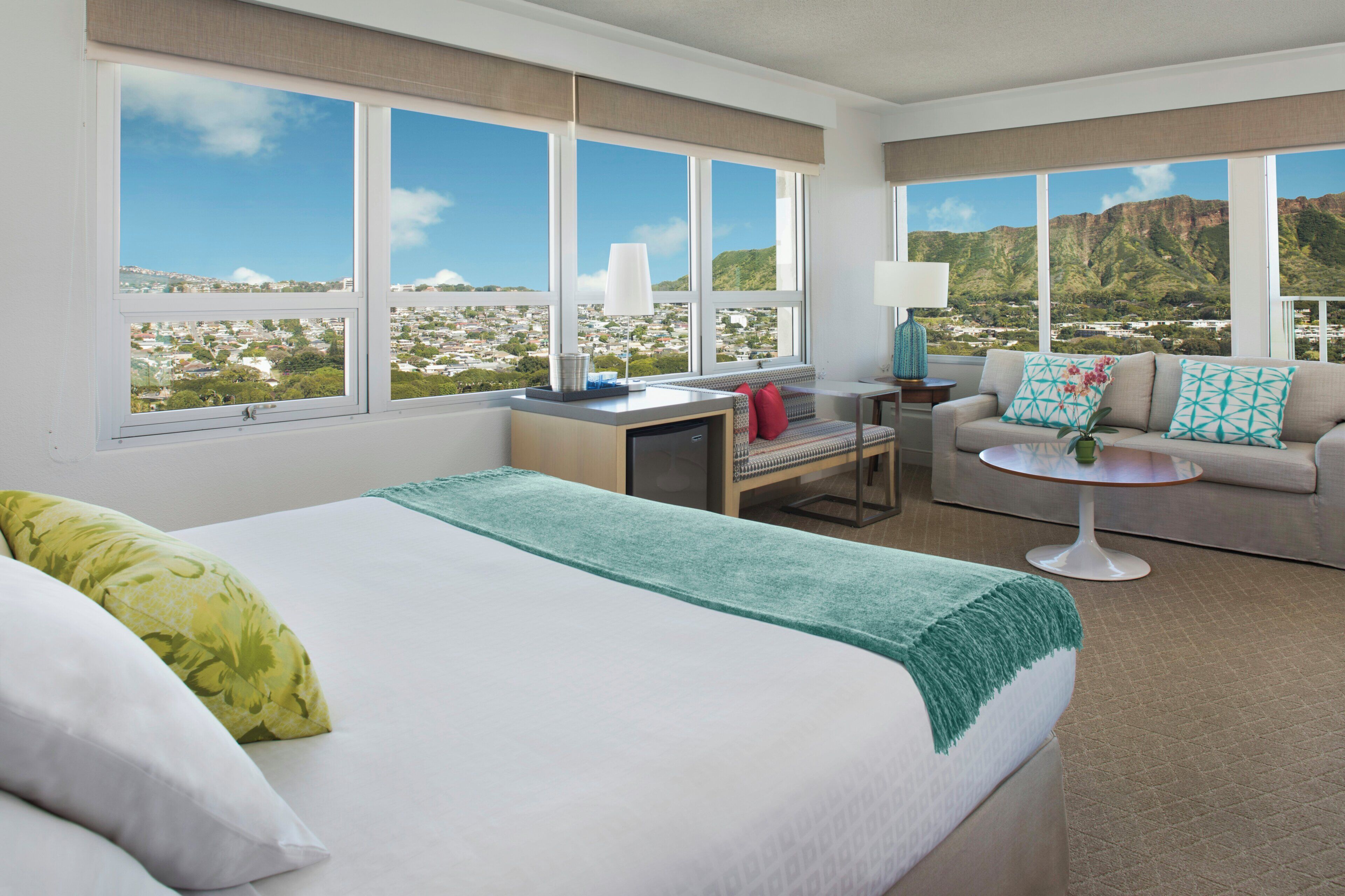 Habitación de hotel con cama, sofá, una mesita y amplias ventanas con vista a la ciudad y la montaña.