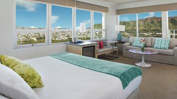 Habitación de hotel con cama, sofá, una mesita y amplias ventanas con vista a la ciudad y la montaña.