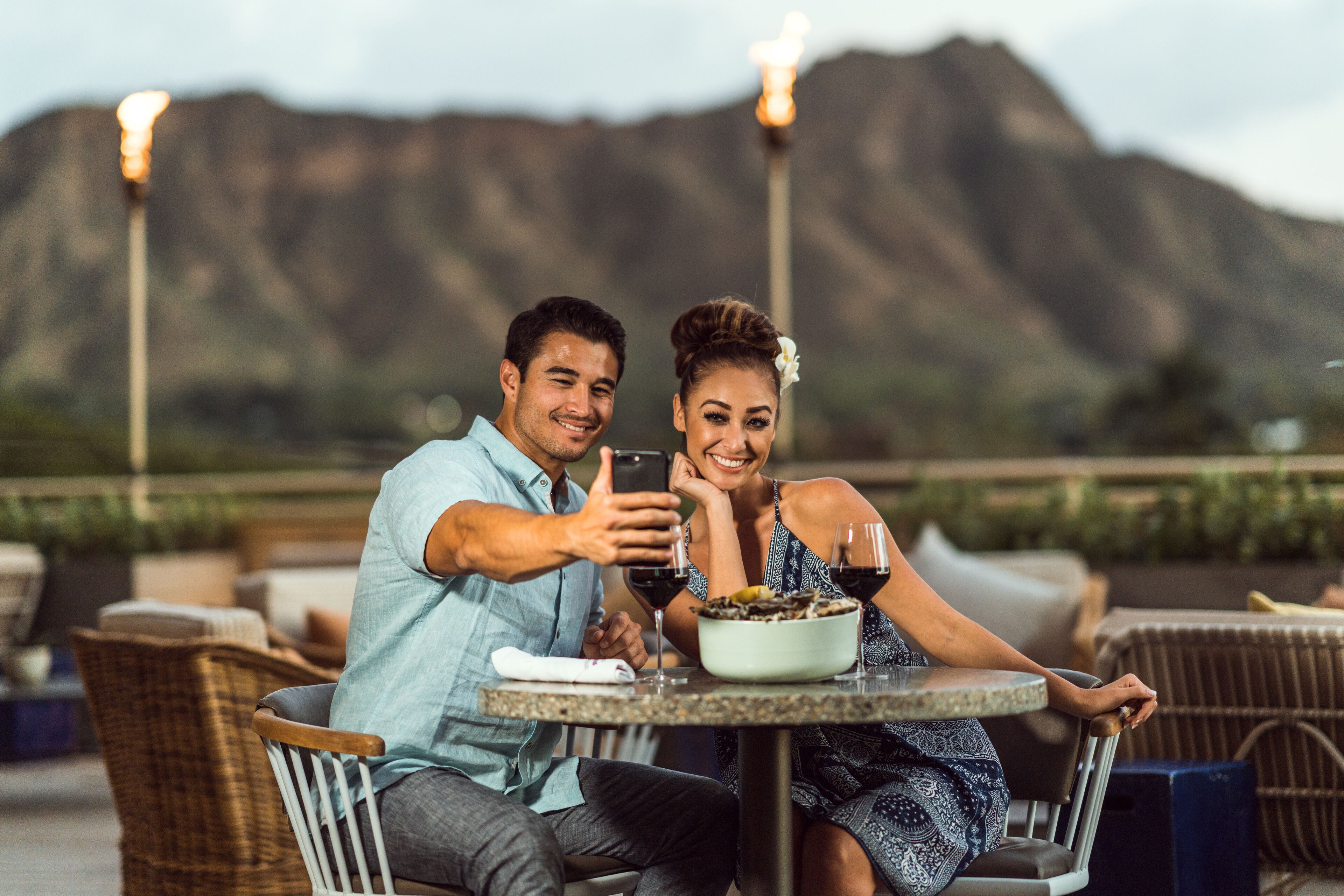 A couple taking a selfie at an outdoor dining area with a mountain view in the background.
