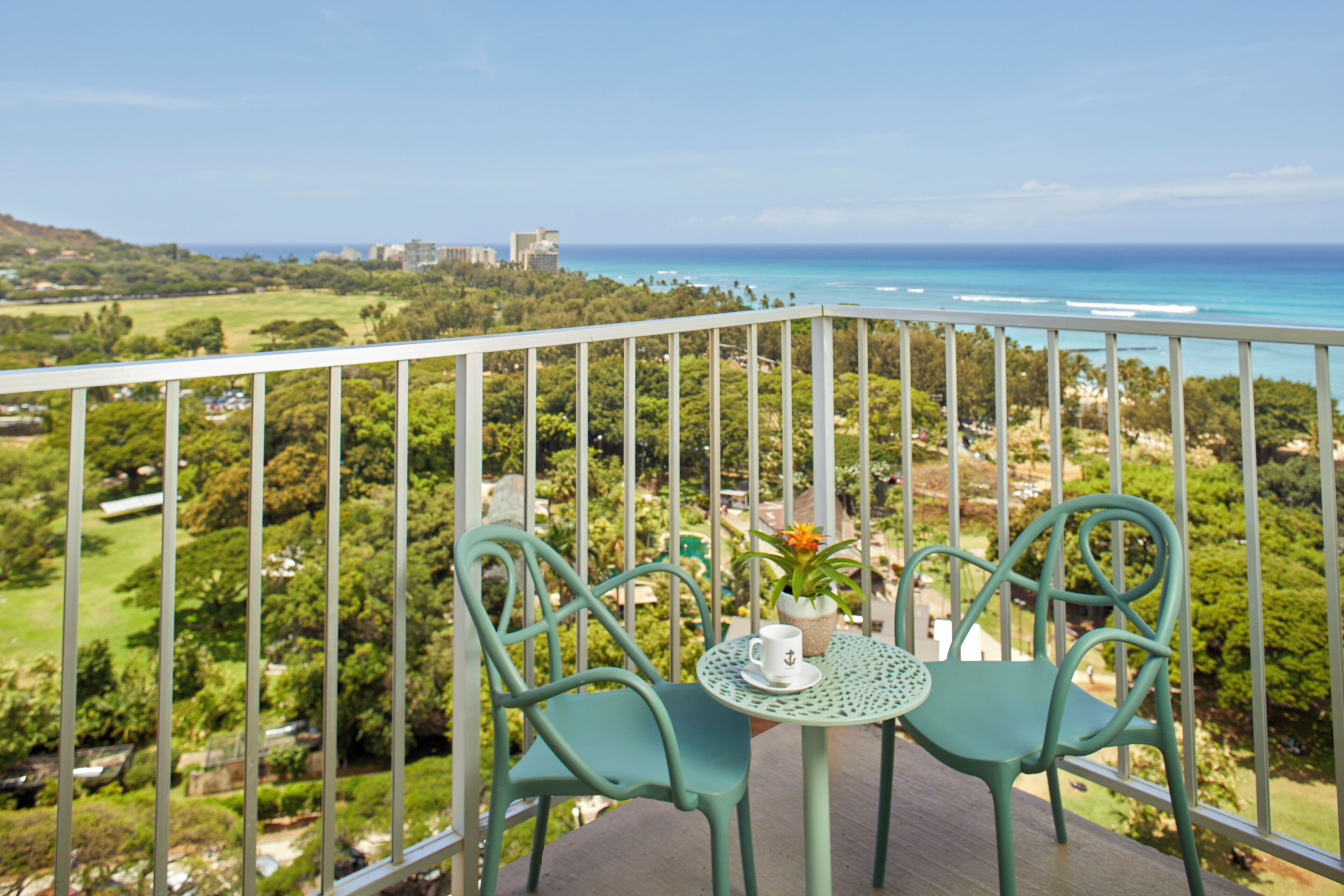 A balcony with two chairs and a table, overlooking a coastal landscape with greenery and buildings.