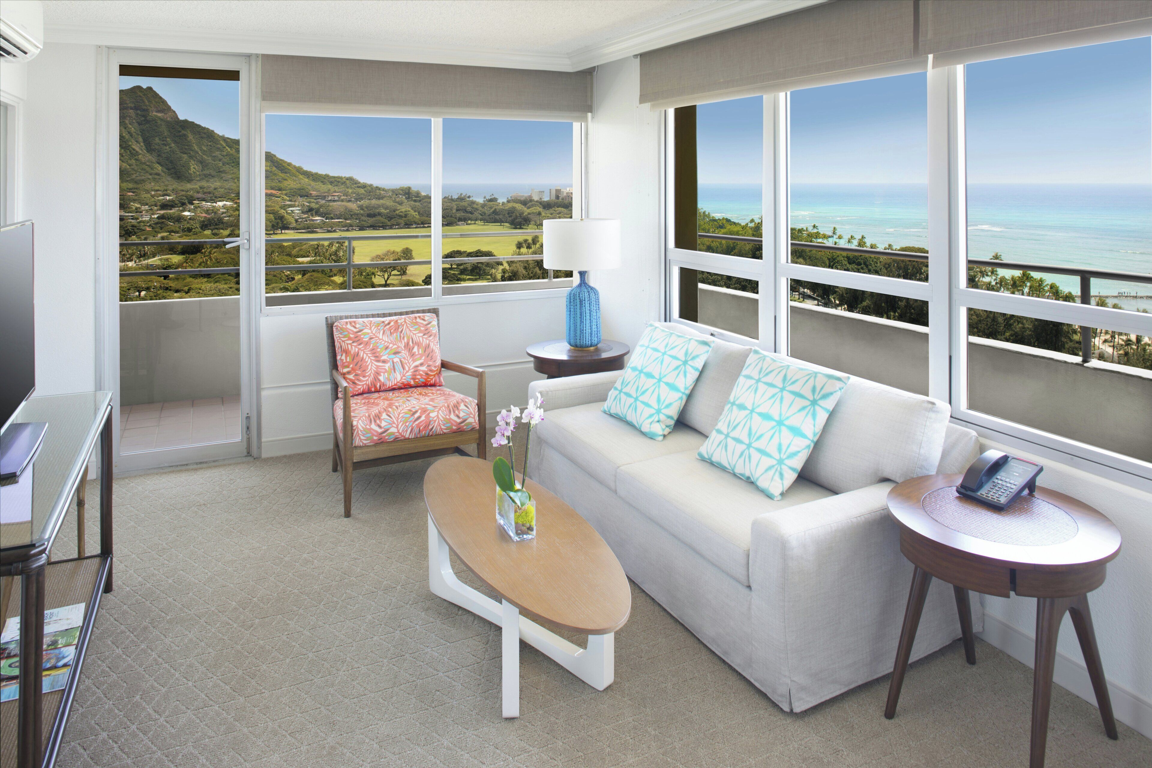 A room with a sofa, armchair, coffee table, and a small side table with a lamp and phone. Large windows offer a view of a mountainous landscape and ocean.