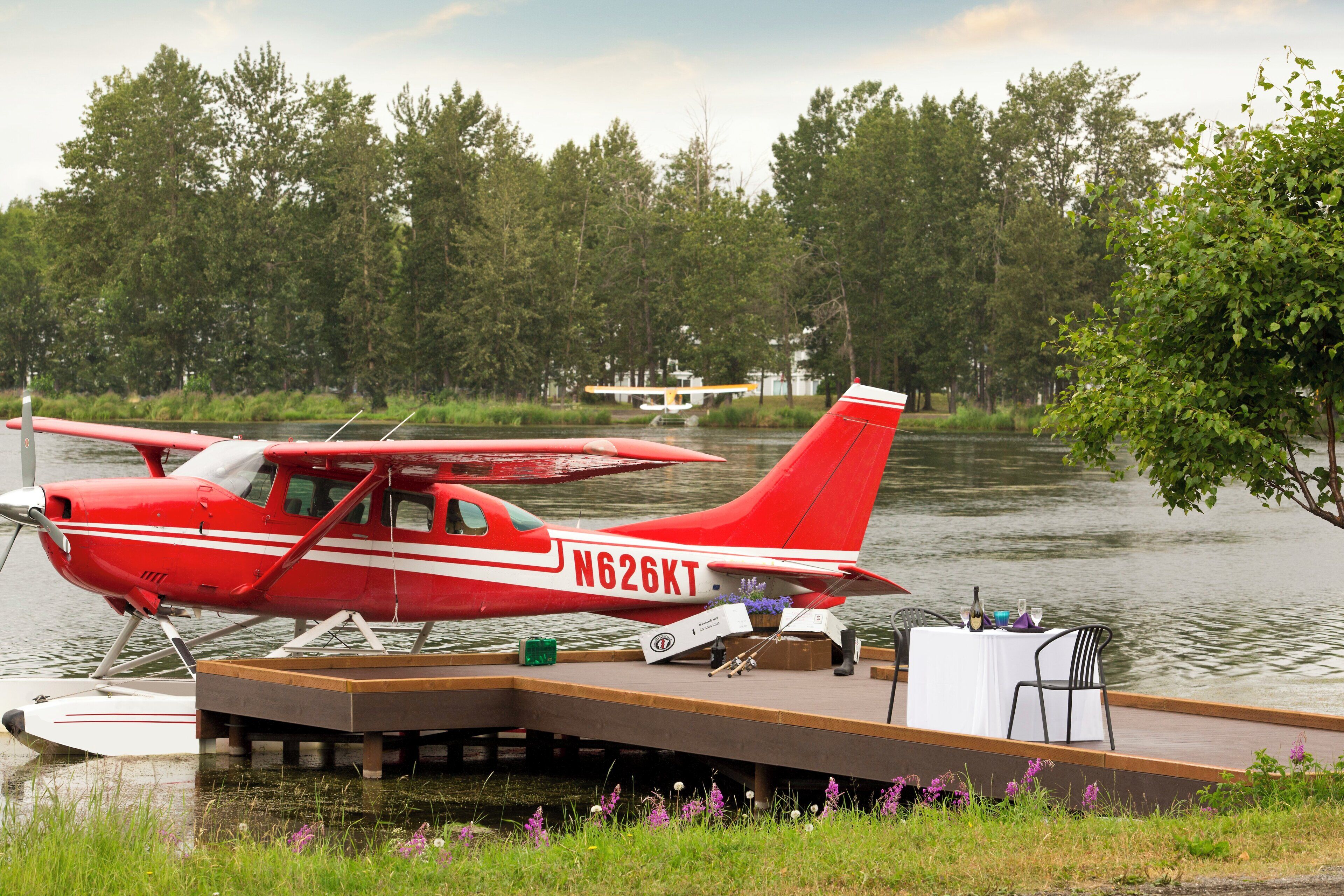 Un hidroavión rojo atracado en un muelle de madera, con una mesa y sillas blancas, rodeado de agua y árboles.