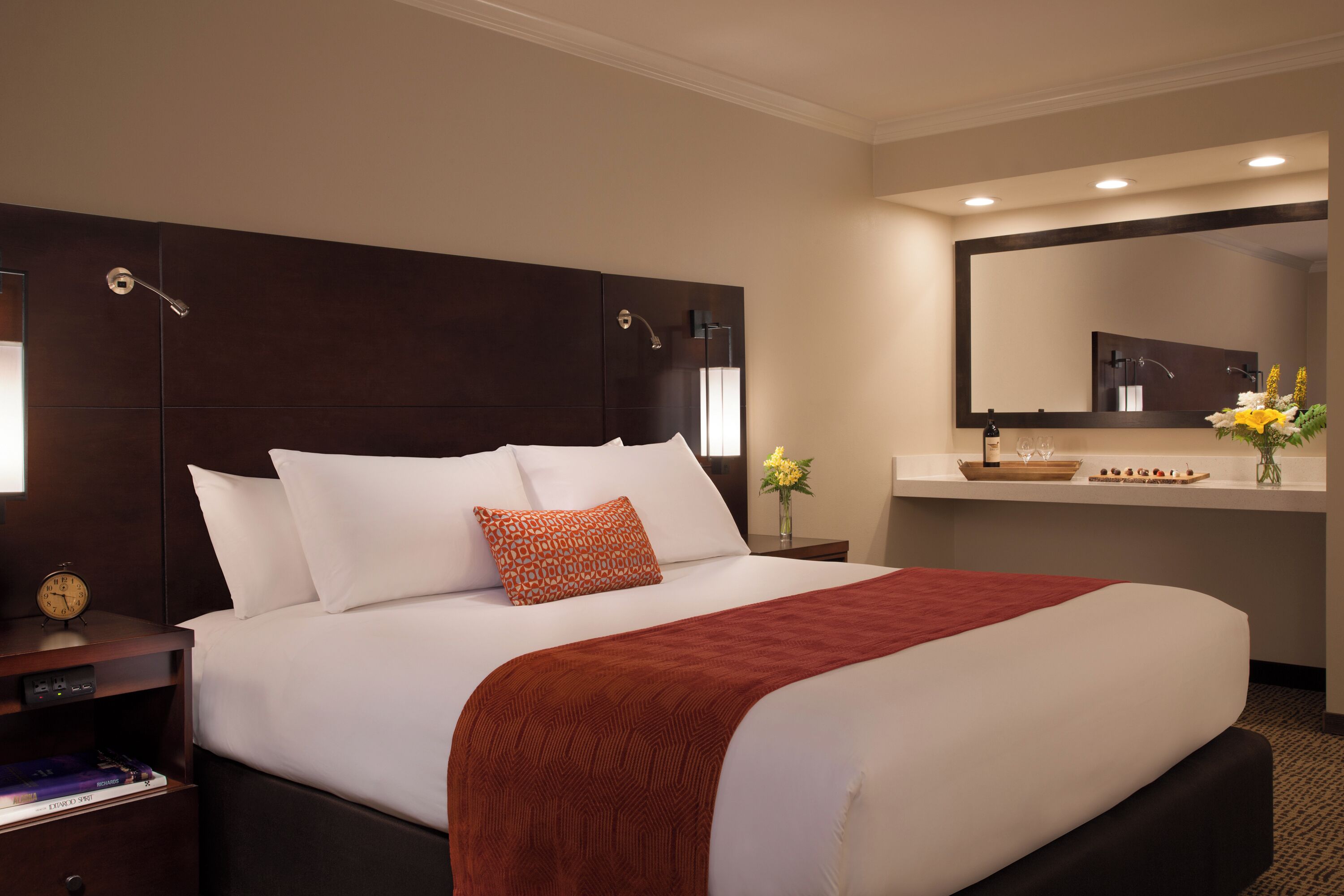 Habitación de hotel con una cama grande, una mesita de noche con un reloj, un tocador con espejo y un jarrón con flores.