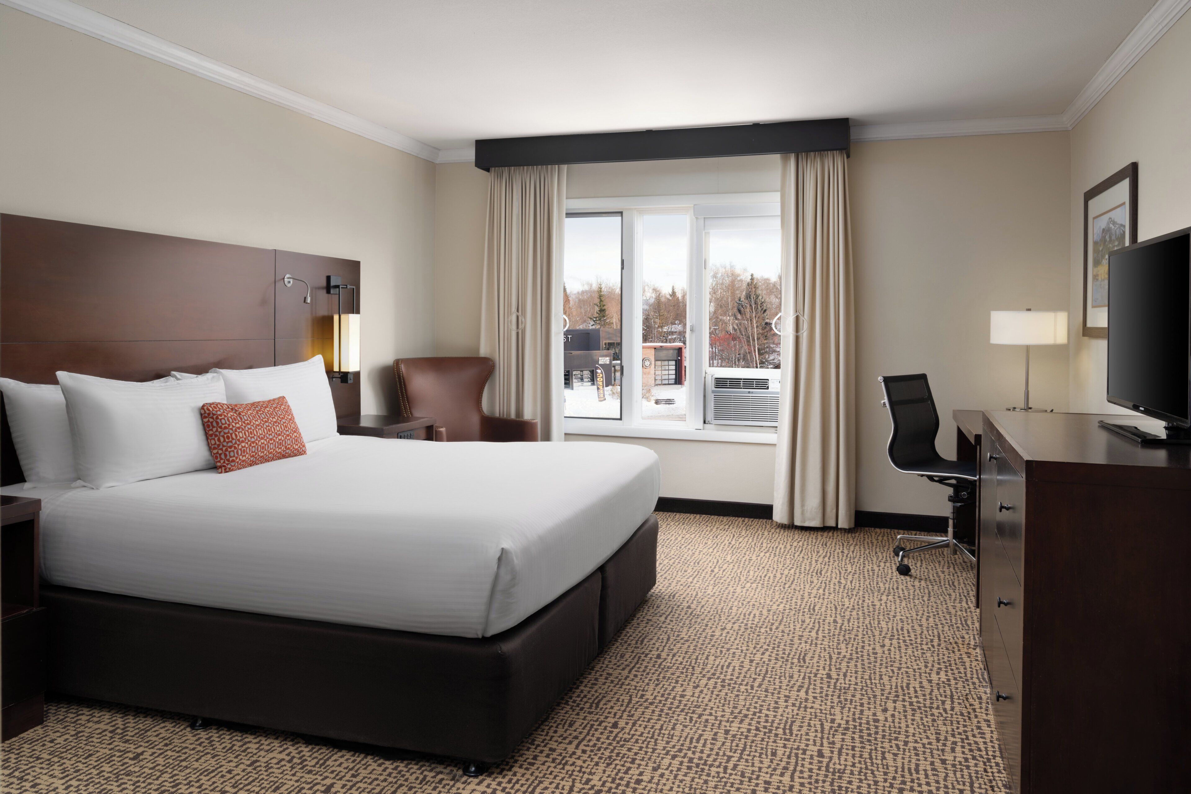 Quarto de hotel com cama grande, escrivaninha, cadeira, televisão e janela com cortinas.