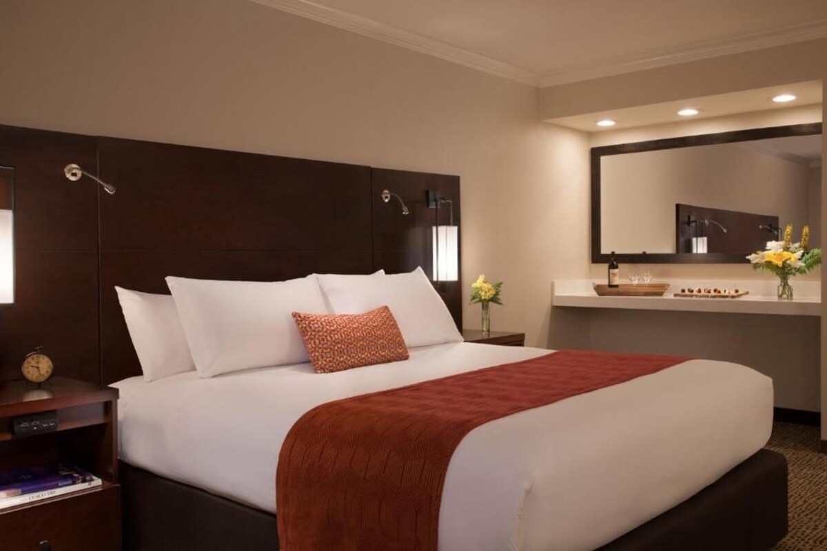 Una habitación de hotel con una cama grande, una mesita de noche, un espejo y un pequeño jarrón con flores.