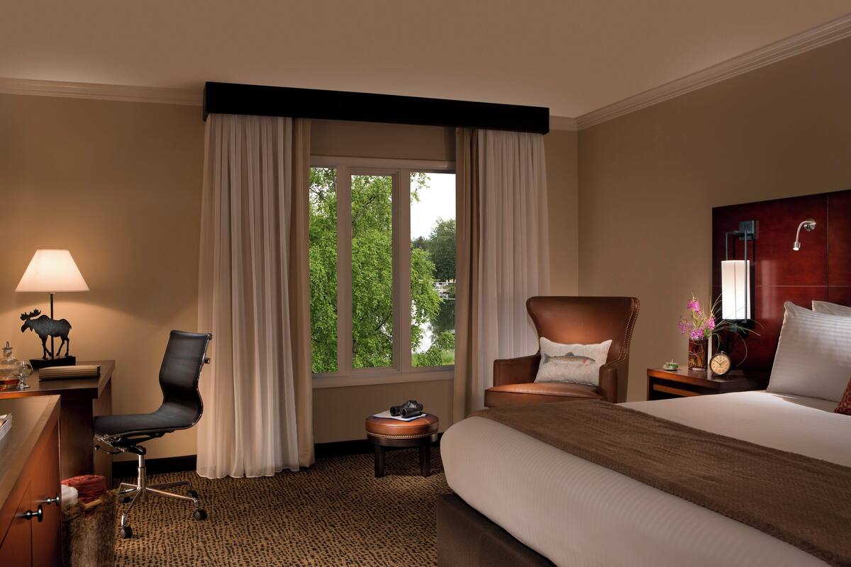 A hotel room with a bed, a desk with a lamp, a chair, a small table with a clock, and a window with curtains.
