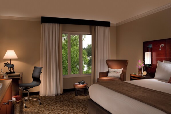 A hotel room with a bed, a desk with a lamp, a chair, a small table with a clock, and a window with curtains.