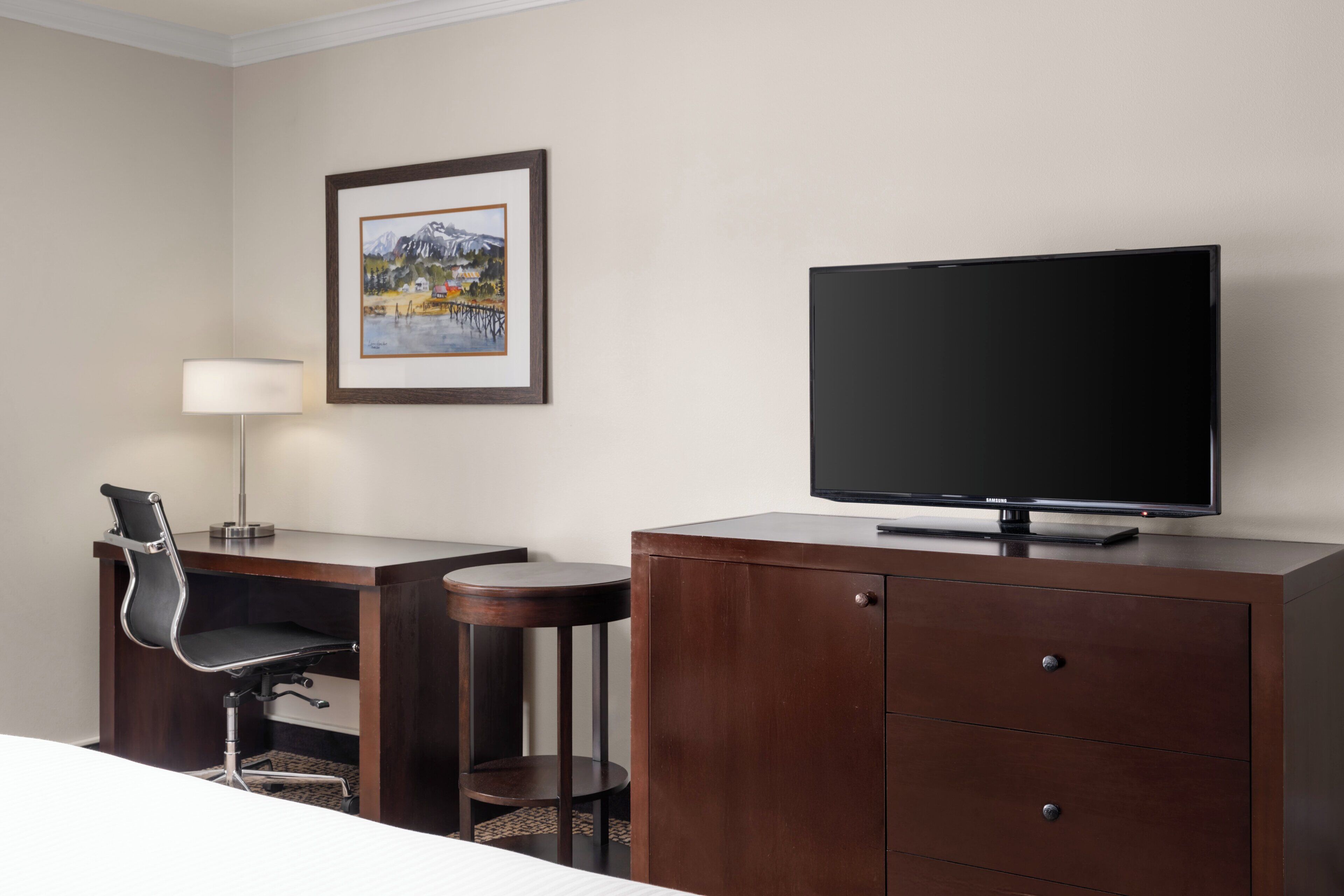 Habitación de hotel con cama, un escritorio con silla, televisor, lámpara y un cuadro en la pared.