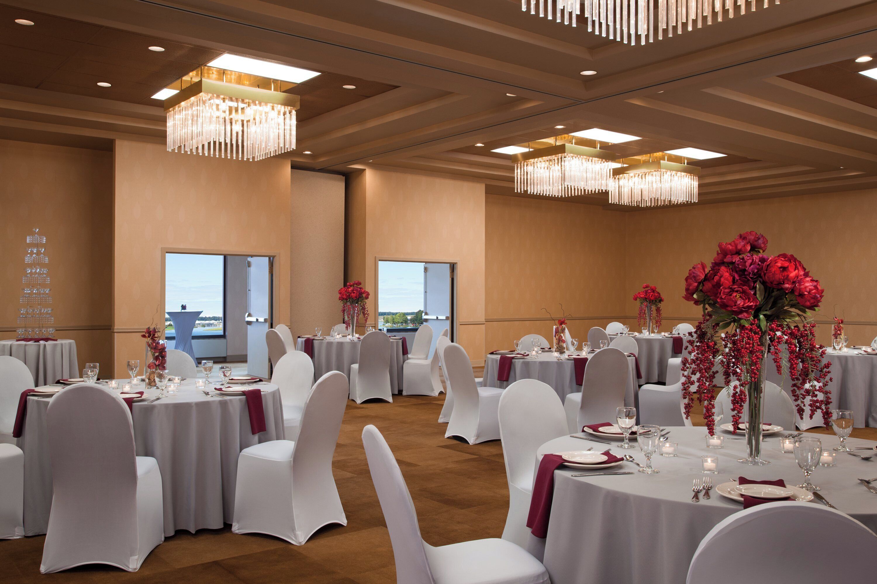 A banquet hall with round tables, white chairs, chandeliers, and floral centerpieces.