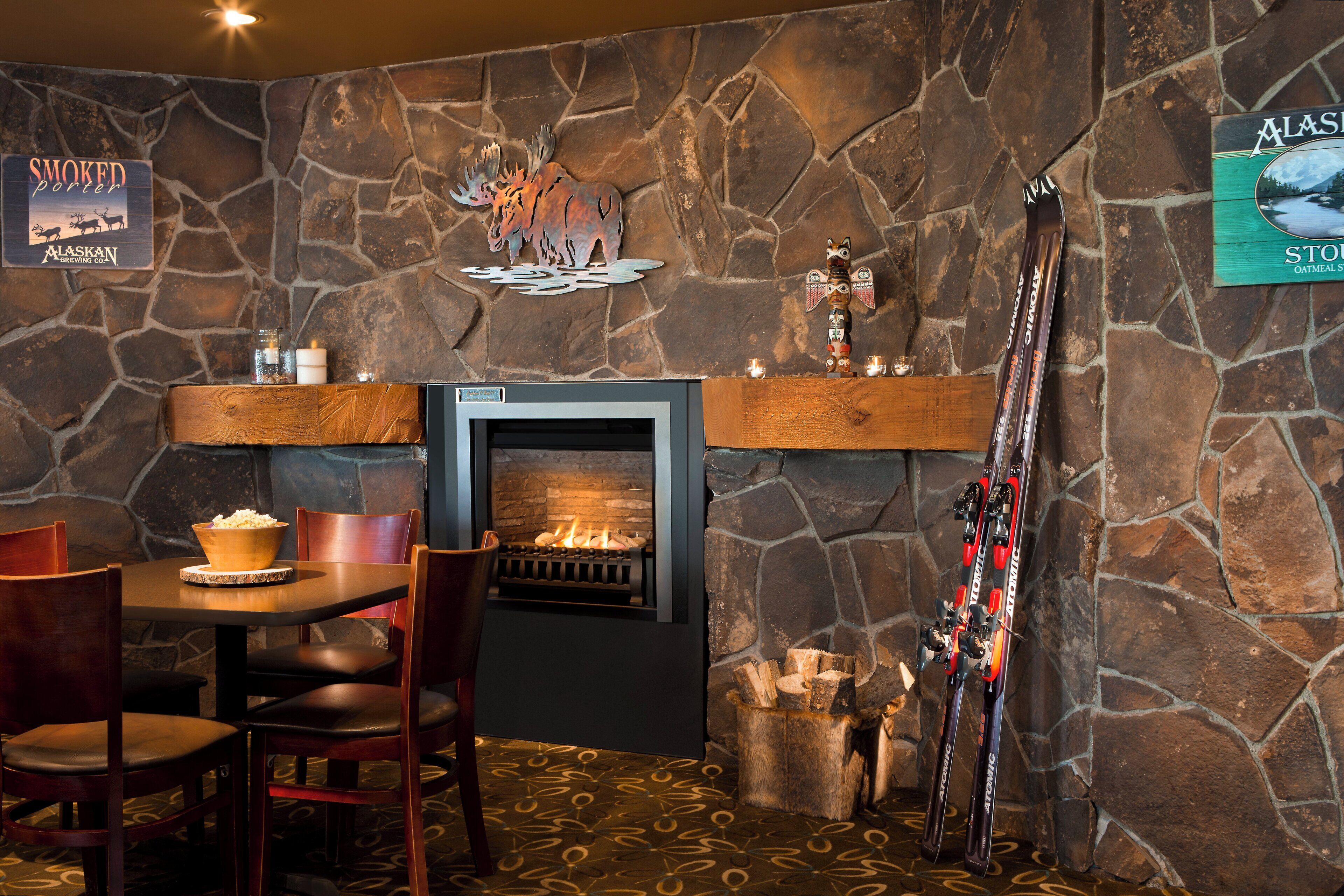 Pared de piedra con chimenea, sillas de madera, una mesa con un tazón de palomitas de maíz y esquíes apoyados en la pared.