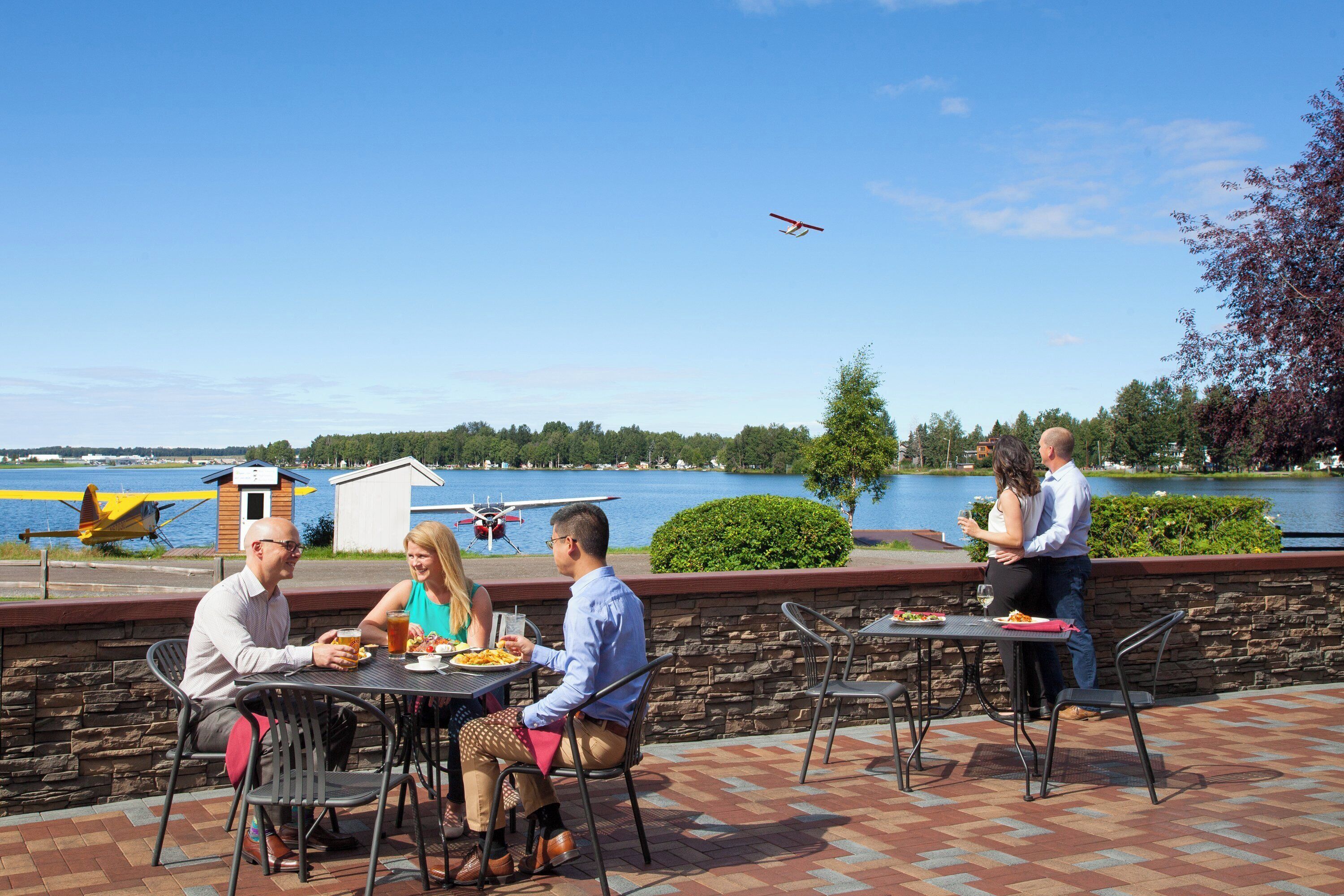 A waterfront patio with tables and chairs, a small airplane display, and a lake view.