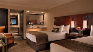 A hotel room with two beds, a sitting area with a chair, a small table with a vase of flowers, and a mirror above the bed.