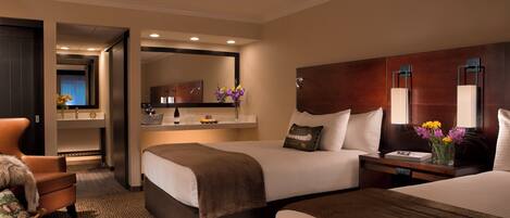 Ein Hotelzimmer mit zwei Betten, einer Sitzecke mit einem Sessel, einem kleinen Tisch mit Blumenvase und einem Spiegel über dem Bett.