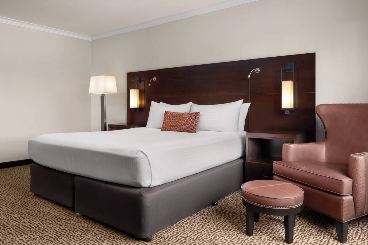 A hotel room with a large bed, a nightstand, a lamp, a chair, and a carpeted floor.