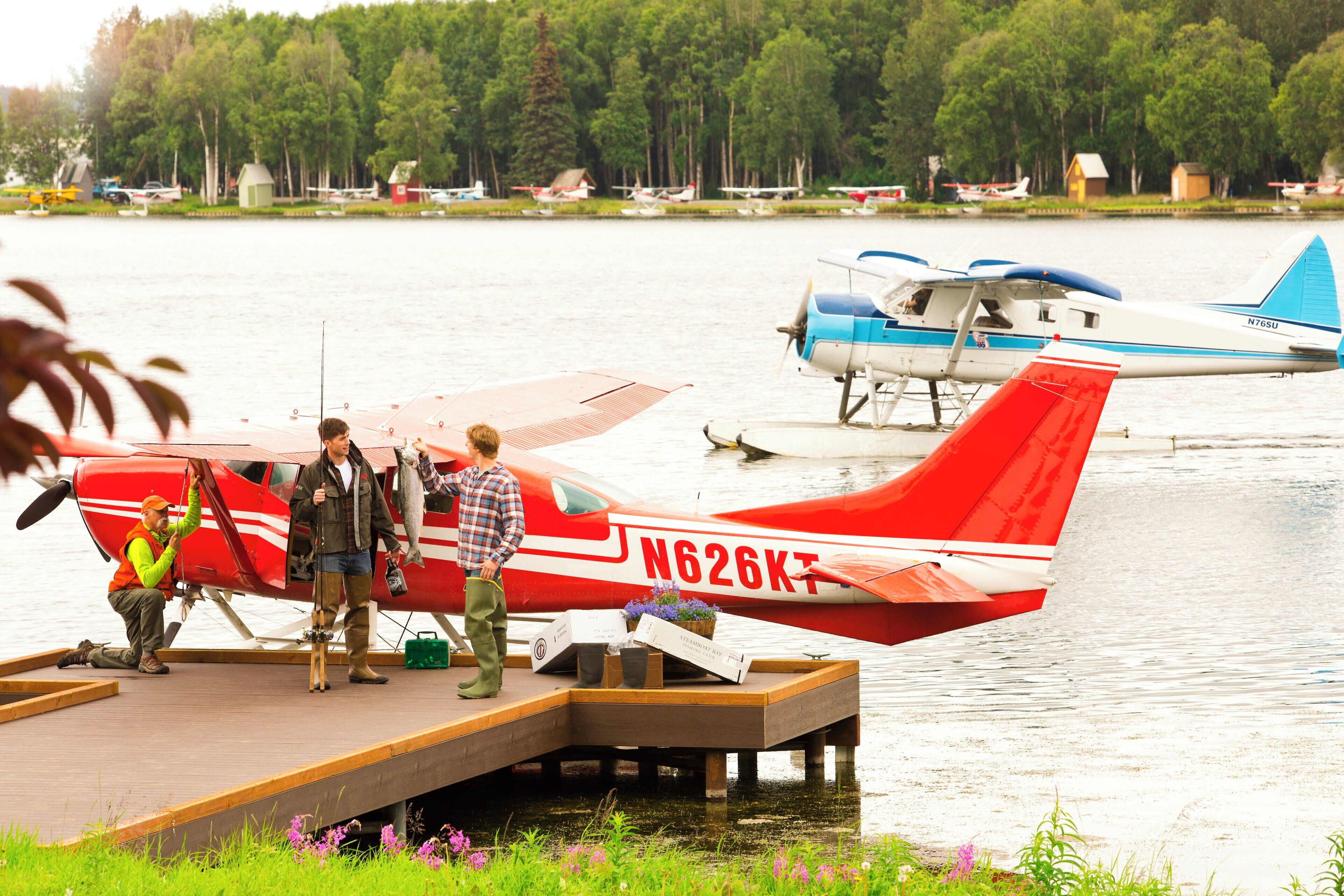 Un petit avion rouge est garé sur un quai en bois, au bord d’un lac, avec d’autres avions en arrière-plan.