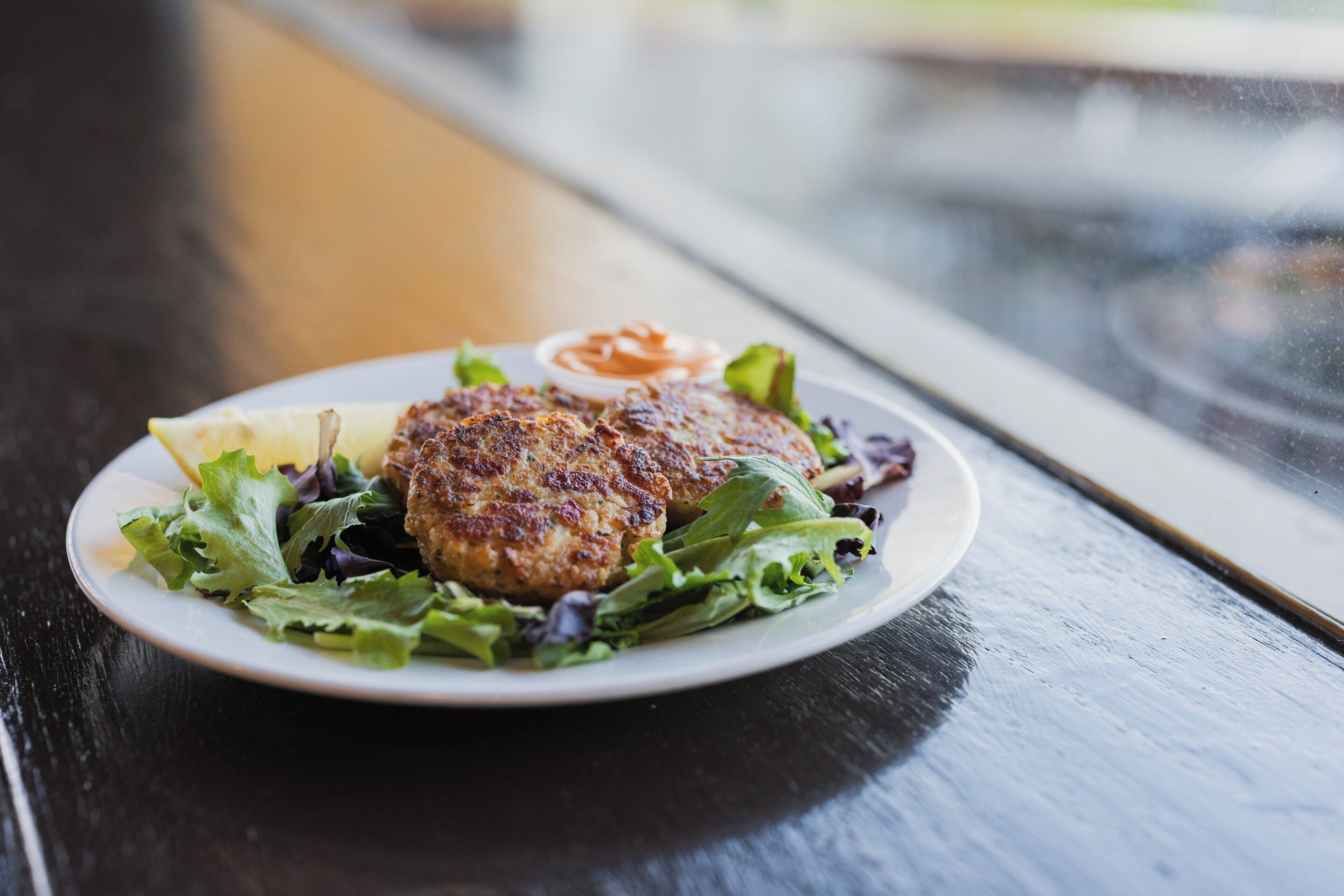 A plate with a burger, salad, and a small bowl of sauce on a table.
