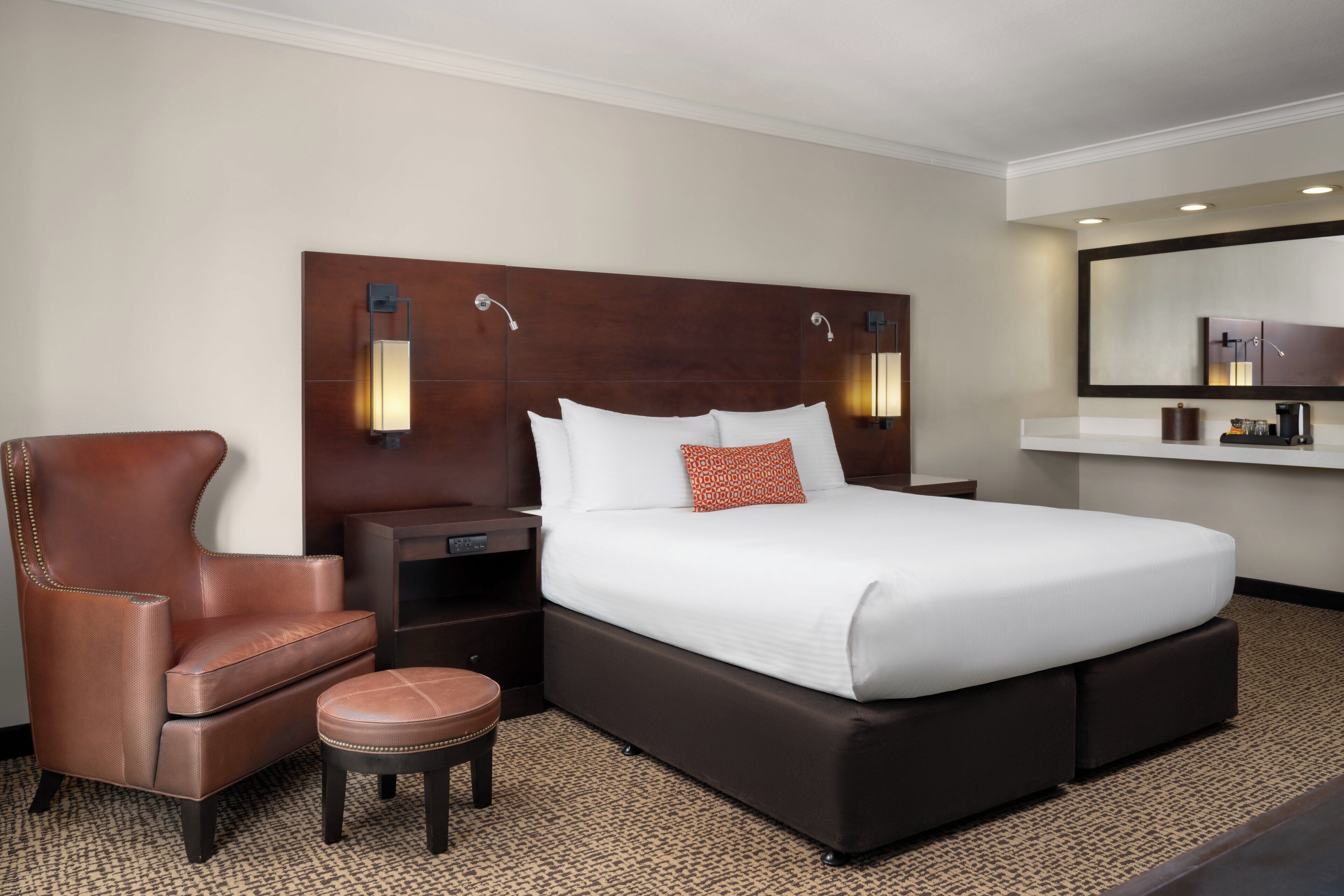 A hotel room with a large bed, a leather armchair, a small stool, a nightstand, and a mirror above the bed.