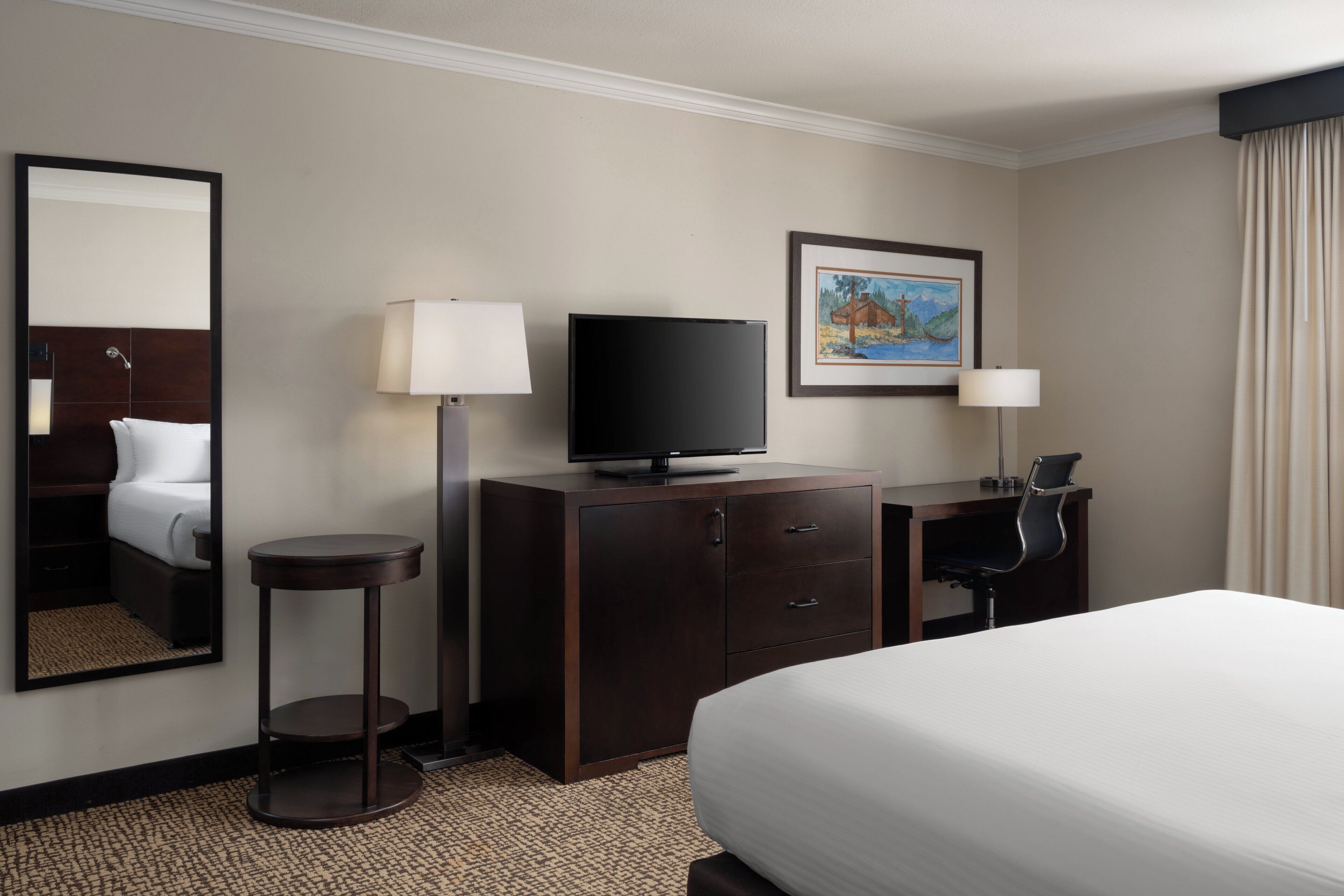 Quarto de hotel com cama, escrivaninha com cadeira, televisão, luminária de mesa, quadro na parede e espelho.