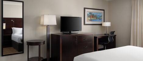 Habitación de hotel con una cama, un escritorio con una silla, una televisión, una lámpara de escritorio, un cuadro en la pared y un espejo.