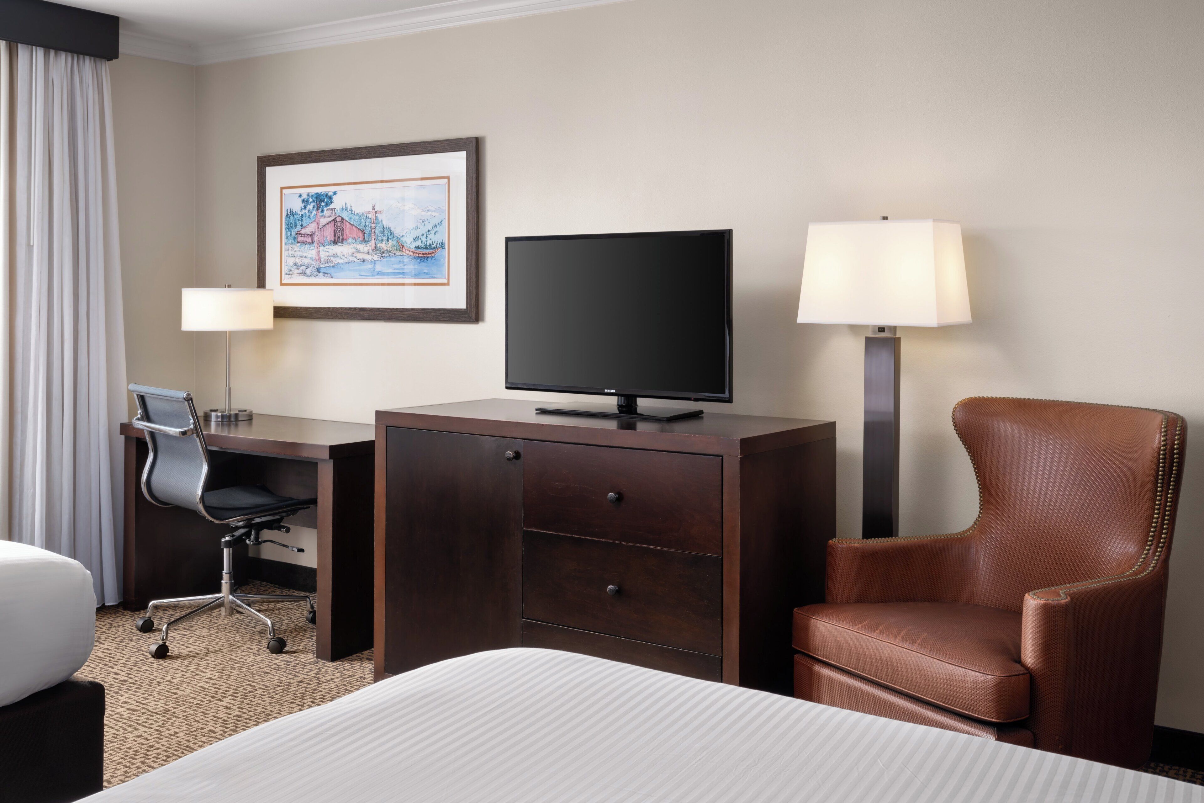 A hotel room with a bed, a desk with a chair, a television, a lamp, and a framed picture on the wall.
