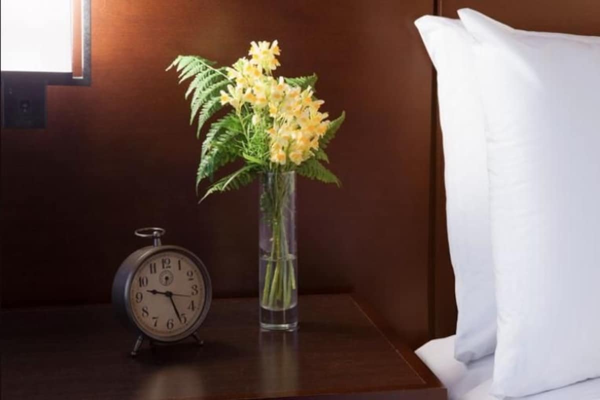 A hotel room with a bed, a white pillow, a nightstand with a clock and a vase of flowers.
