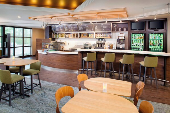 Un área de bar moderna con mesas y sillas de madera, una barra con carta y una estantería llena de botellas.