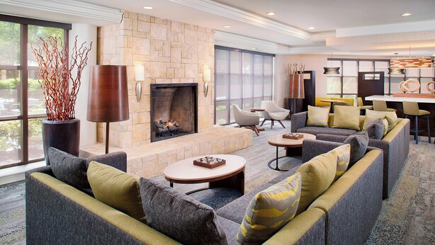 A modern living room with a stone fireplace, comfortable seating, and large windows offering a view of the outdoors. - Courtyard By Marriott Ann Arbor (Ann Arbor)