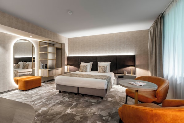 Una habitación de hotel moderna con una cama grande, un espacio de descanso con una mesa redonda y una estantería empotrada.