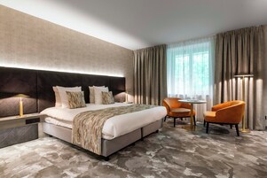 Une chambre d’hôtel moderne avec un grand lit, une table de chevet avec une lampe, un fauteuil orange, une petite table ronde et une vue sur la verdure à travers des rideaux transparents.