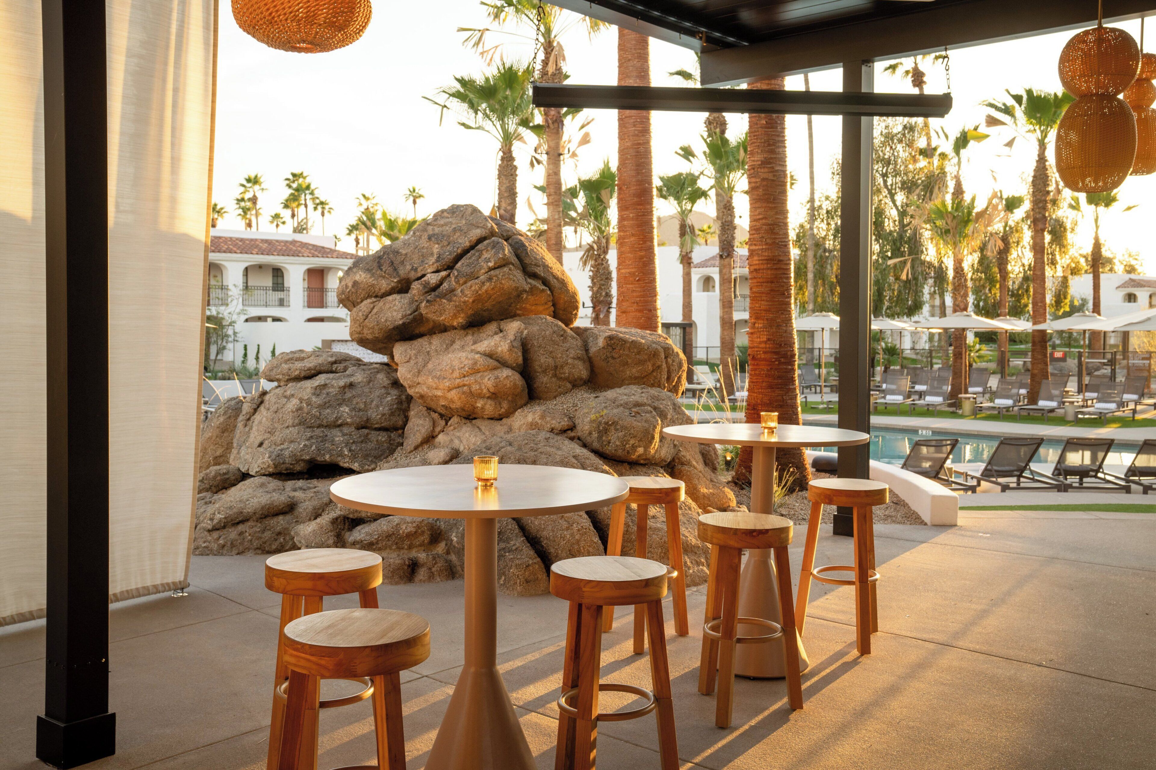 A patio area with round tables and stools, a rock formation, and a pool in the background.