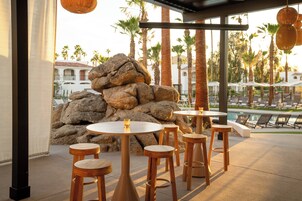 A patio area with round tables and stools, a rock formation, and a pool in the background.